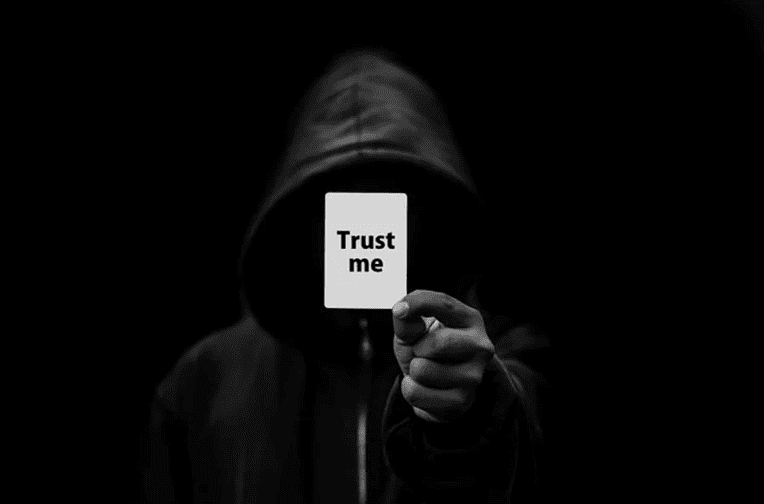 Guy wearing black cape with black background holding a white paper with 'trust me' 